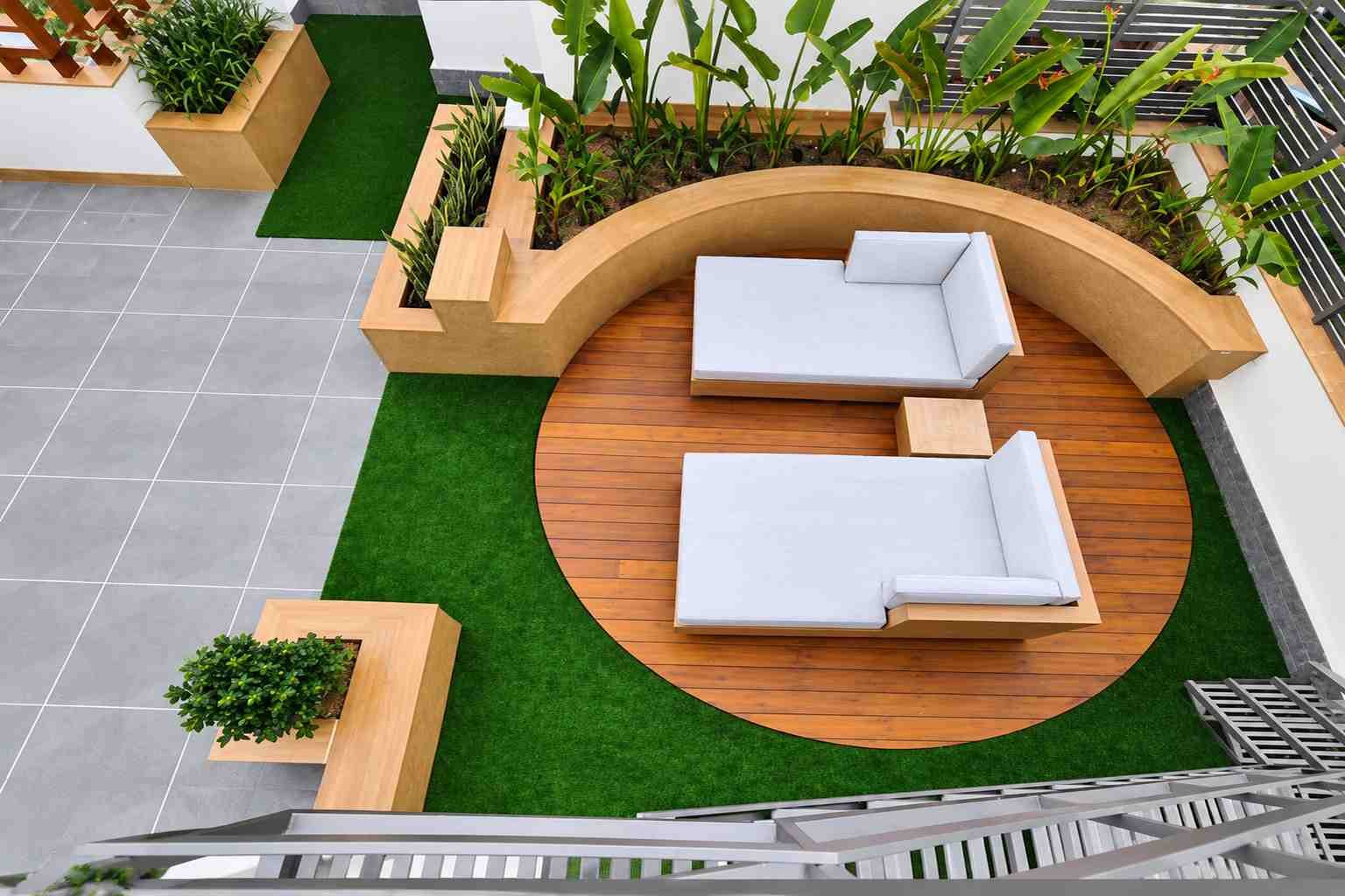 Modern rooftop outdoor lounge with a circular wooden deck, two white lounge sofas, curved planter seating, lush tropical plants, green turf, and contemporary flooring.