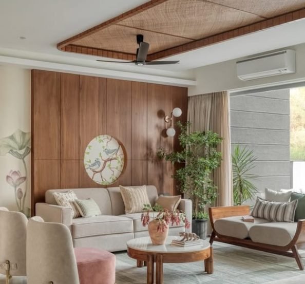Modern luxury living room with a warm wooden accent wall, beige sofa, round wooden coffee table, indoor plants, and a stylish false ceiling with a ceiling fan.